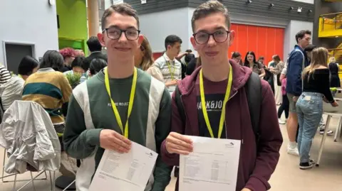 BBC Identical twins, both with glasses, one wearing a green jumper and one in a red jacket. Both are holding up their A-level results. 