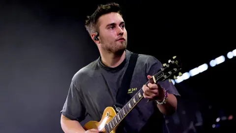 Getty Images Sam Fender looks to his left as he plays a yellow electric guitar. He is wearing a dark t-shirt and has brown short hair and brown stubble. There is a dark black background.