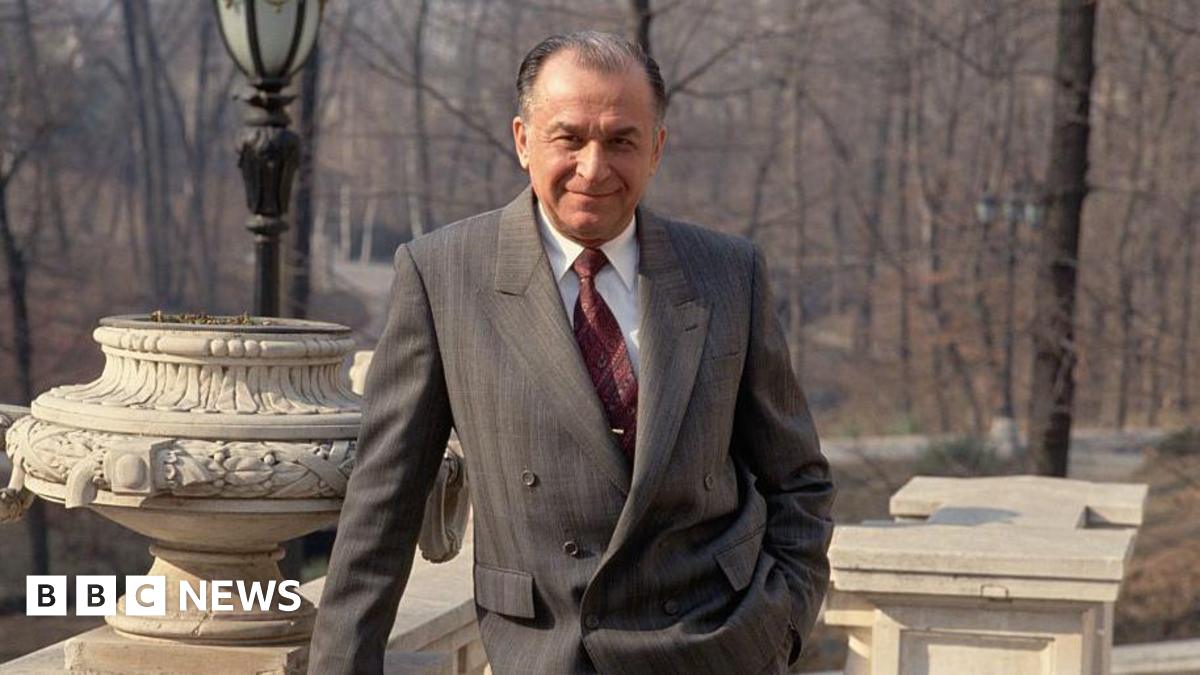 Iliescu poses and looks straight down the camera with a slight smile. He is leaning on a stone balcony and wearing a grey suit.