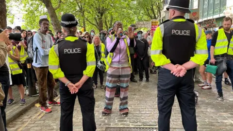 BBC Police with their backs to the camera holding a line in front of a large number of counter-protesters. One woman is shouting into a megaphone in front of them. 