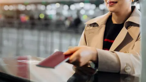Getty Images A woman with pink lipstick wearing a stone coloured trench coat and black jumper with red stripes passing over her passport as she stands at a desk in what appears to be an airport. The background is blurred.