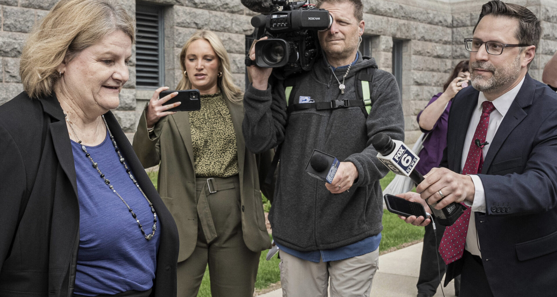 A woman is approached by reporters and a camera crew outside a stone building; microphones and cameras are directed toward her as she walks.
