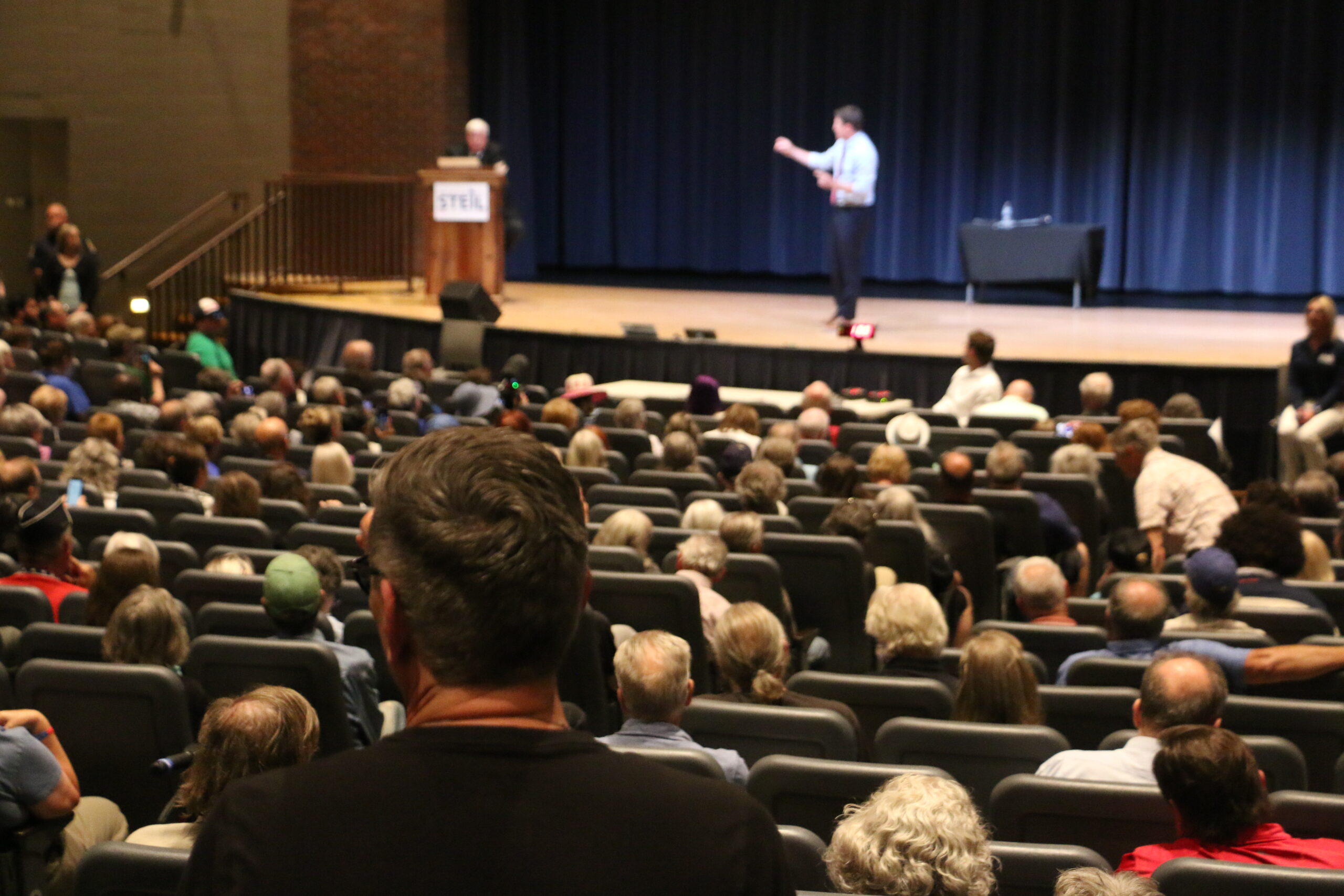 A large audience seated in an auditorium listens to two speakers on stage, one standing at a podium and the other gesturing while speaking.