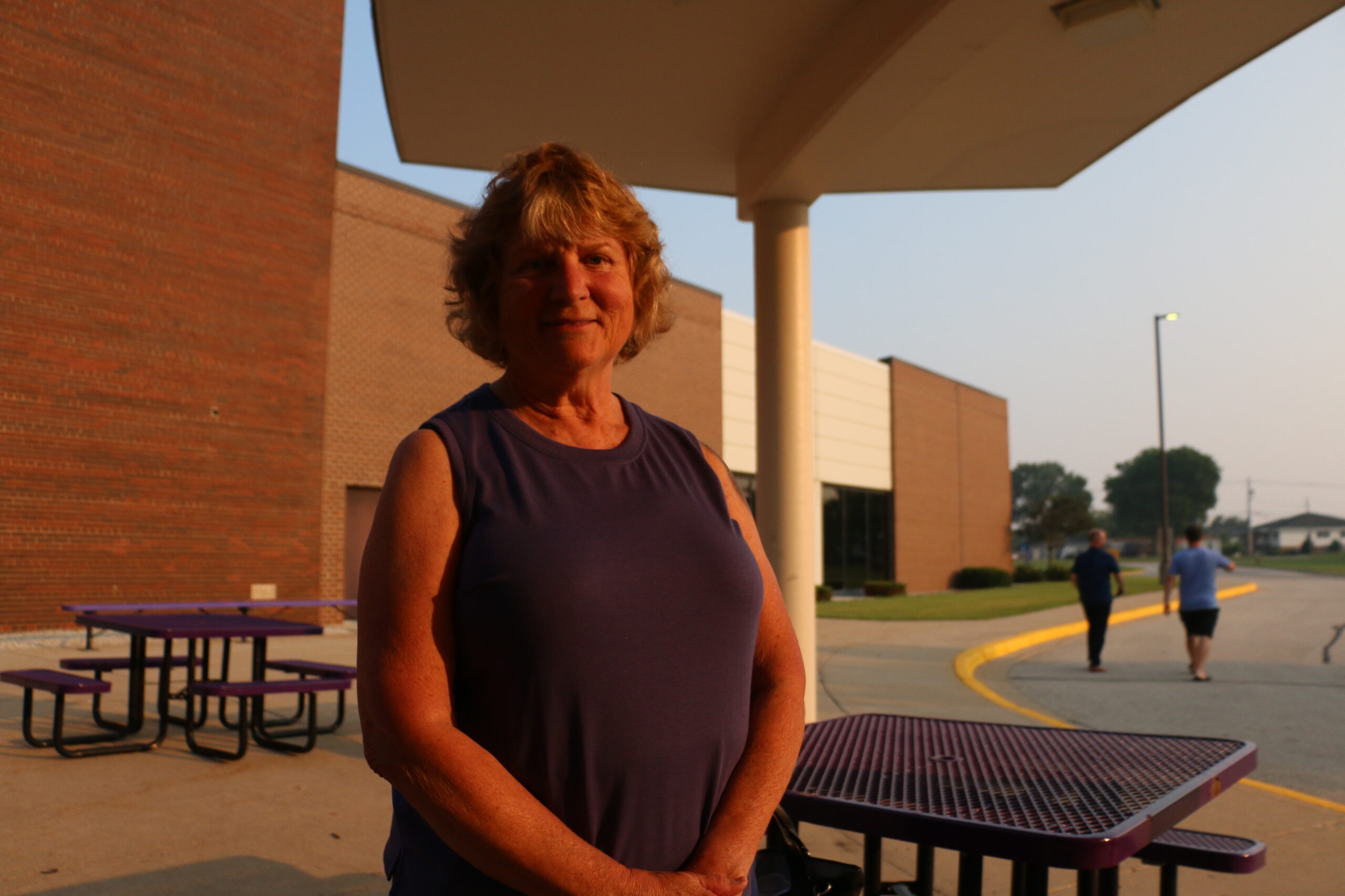 A woman in a sleeveless purple top stands in front of a brick building near outdoor tables, with two people walking in the background.