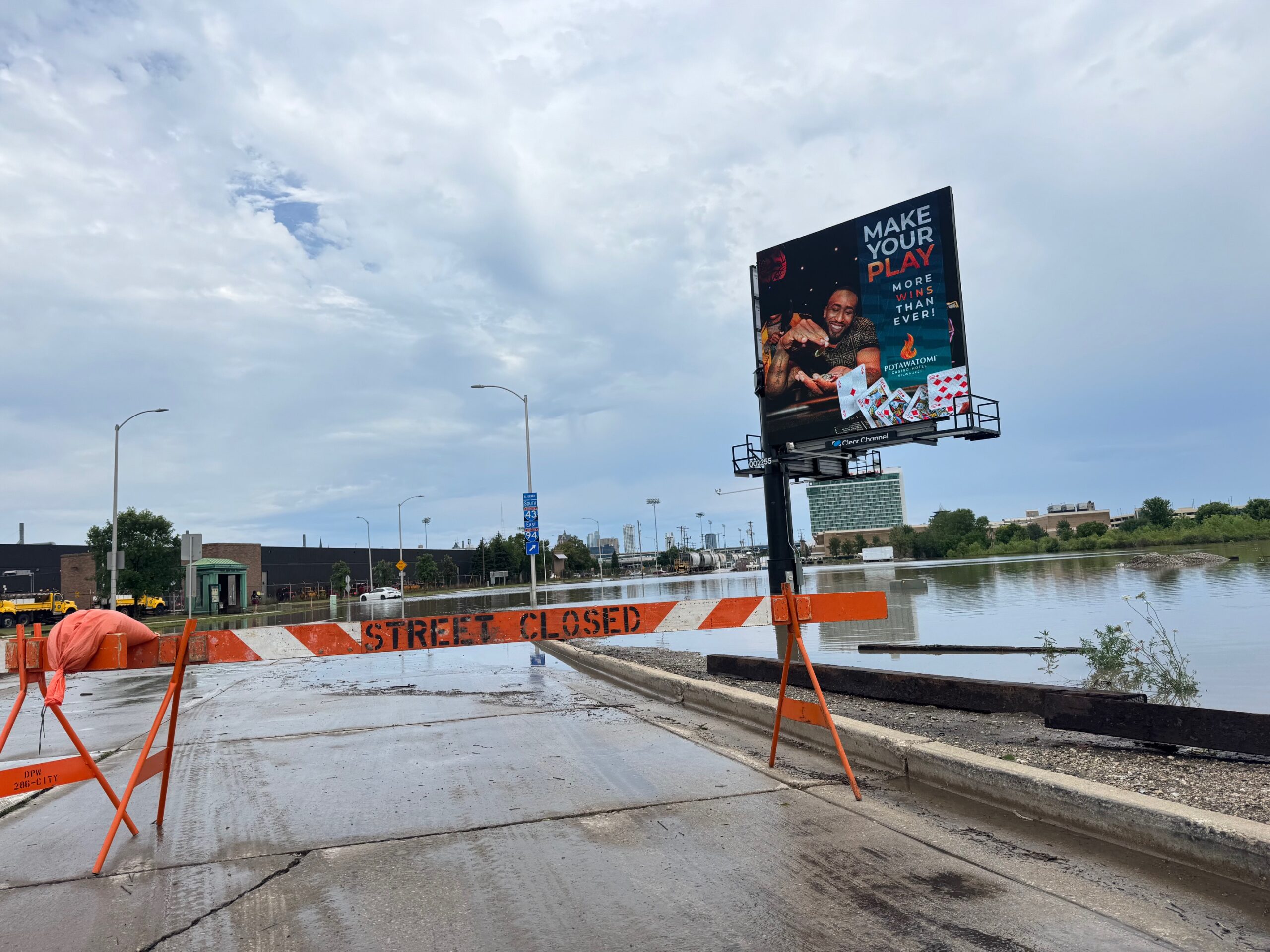 A Street Closed barrier blocks a road near a flooded area, with a casino advertisement billboard and cloudy sky in the background.