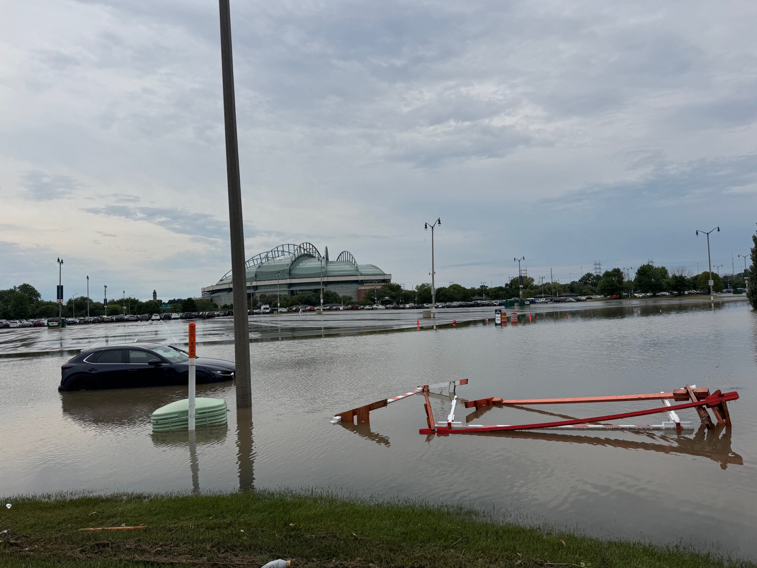 A flooded parking lot with a partially submerged car, floating debris, and a large stadium visible in the background under a cloudy sky.