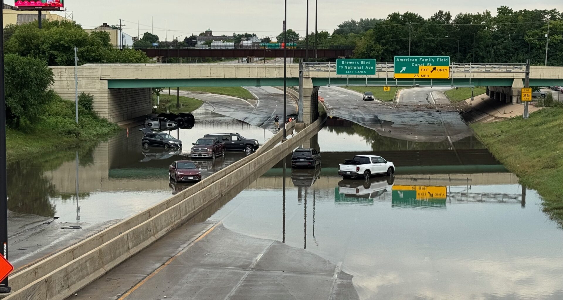 Several vehicles are stranded on a flooded highway under an overpass, with water covering multiple lanes and highway signs visible above.