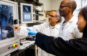 Mak Paranjape in a lab coat with two students looking at a lab device