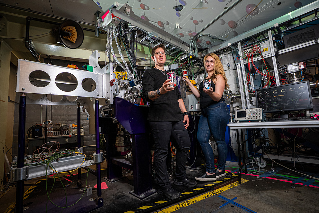 Two people in safety glasses hold glass beakers containing ball-and-stick models of nobelium molecules. They are surrounded by lab equipment that includes wires, foil-wrapped cables, metal apparatuses, and a computer screen showing data. Images of molecules are projected on the ceiling panels.
