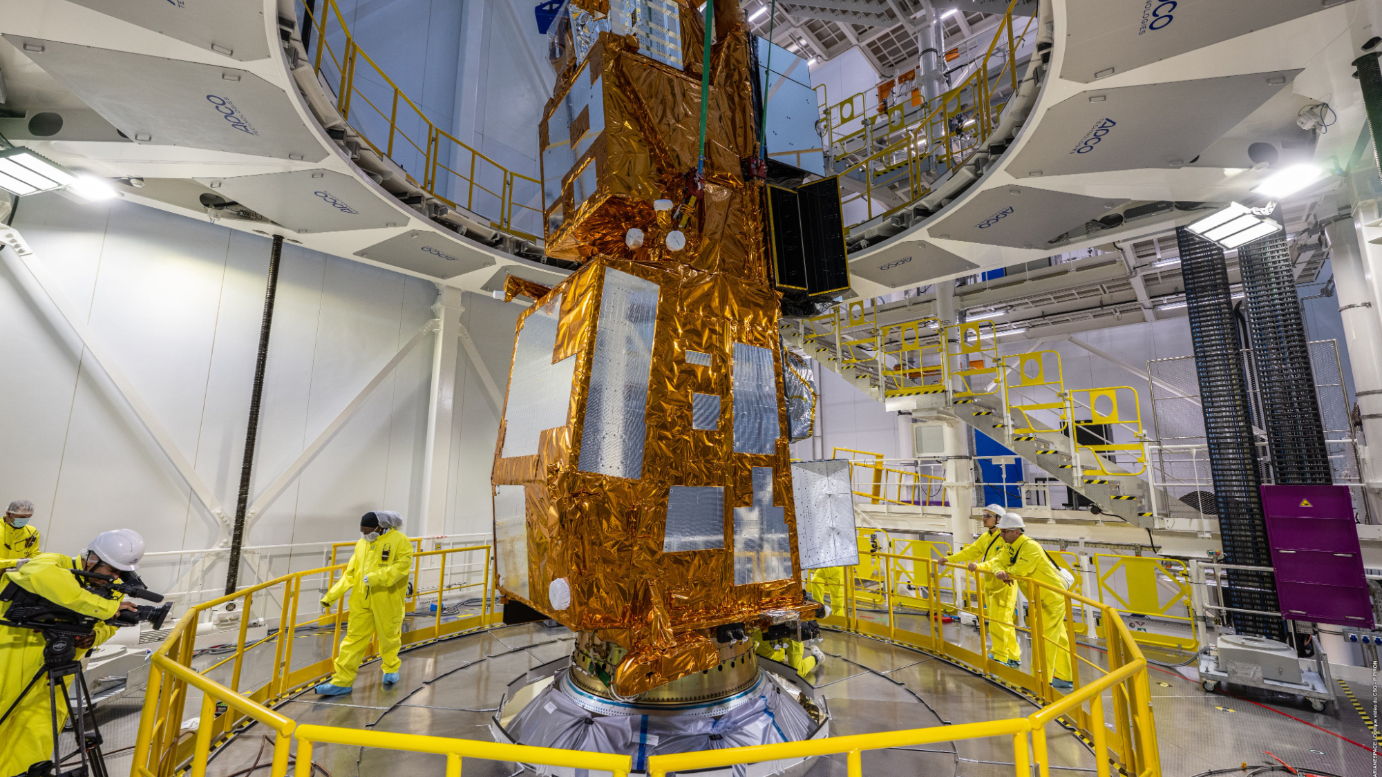a boxy spacecraft wrapped in gold foil is seen inside a large, white-walled clean room