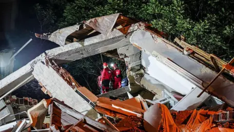 Sergen Sezgin/Anadolu via Getty Images Emergency workers can be seen through a square frame that is being created by a massive chunk of collapsed building.