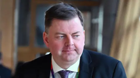 Getty Images Colin Smyth, a man with flat brown hair, while wearing a suit, white shirt and purple tie and multicoloured lanyard