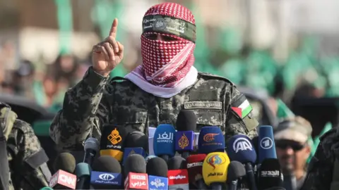 Getty Images Abu Obeida speaks behind a number of microphones with logos of world media while wearing a scarf over his face, flanked by two Hamas fighters.