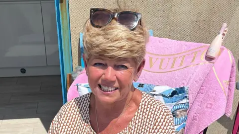 BBC Debbie Booker is sitting outdoors. She has short blonde hair and is wearing sunglasses on top of her head and a patterned shirt. Behind her, a pink towel with the word "VUITTON" is draped over a chair. A bottle is placed on the chair's armrest. The background shows part of an open door and tiled flooring.