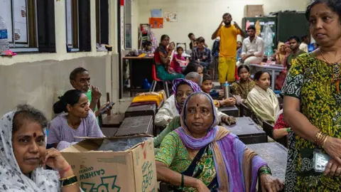 Hindustan Times via Getty Images Women sitting on benches inside a classroom, which has been converted to a temporary shelter. A cardboard box is lying next to one of the women, who is wearing a green saree, and is in the centre of the picture. 