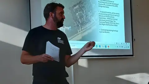 Rufus Pickles Lawrence Bates stands in a meeting room in front of a projector screen. He is looking away from the camera and holds a notepad in one hand, while his other hand is slightly reaching forward as though he is trying to emphasise his point. Mr Bates has short black hair and a short black beard. He is wearing dark trousers a short sleeved cotton t-shirt which is black and has the Wildheart Animal Sanctuary logo on the chest. 