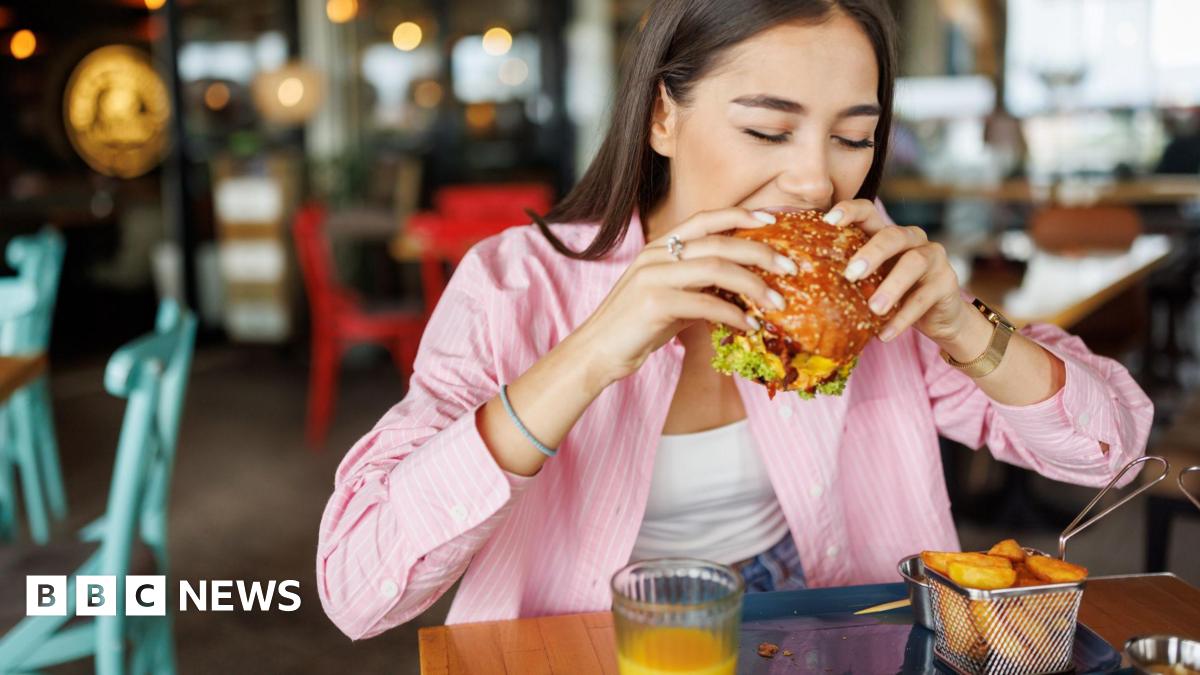 A woman in a pink shirt bites into a large burger inside a casual restaurant.