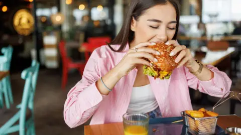 Getty Images A woman in a pink shirt bites into a large burger inside a casual restaurant.