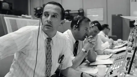 NASA Black and white image of Charlie Duke, along with Jim Lovell and Fred Haise in Nasa Mission Control, during the Apollo 11 mission. They are all dressed in shirts and ties and wear headsets, as they sit in a long line in front of control panels and display screens.