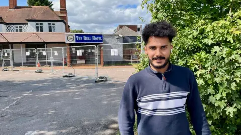 Wael, a man with curly hair, is wearing a navy blue jumper.  He stands in front of fences outside the Bell hotel in Epping which has temporary metal fences outside.