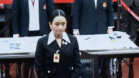 Getty Images A monitor shows Paetongtarn Shinawatra during proceedings at the Constitutional Court in Bangkok on August 21, 2025. She looks glum and is wearing a black suit.    