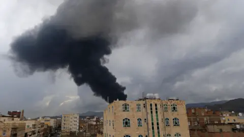 MOHAMMED HUWAIS/AFP via Getty Images A large plume of black smoke rising behind a red-bricked building in Sanaa, one of may including blocks of flats