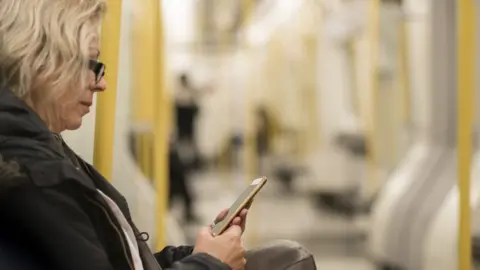 Lovro77/Getty Images A woman with blonde hair in a black coat sits on the Tube and looks at her phone. The carriage is largely empty.