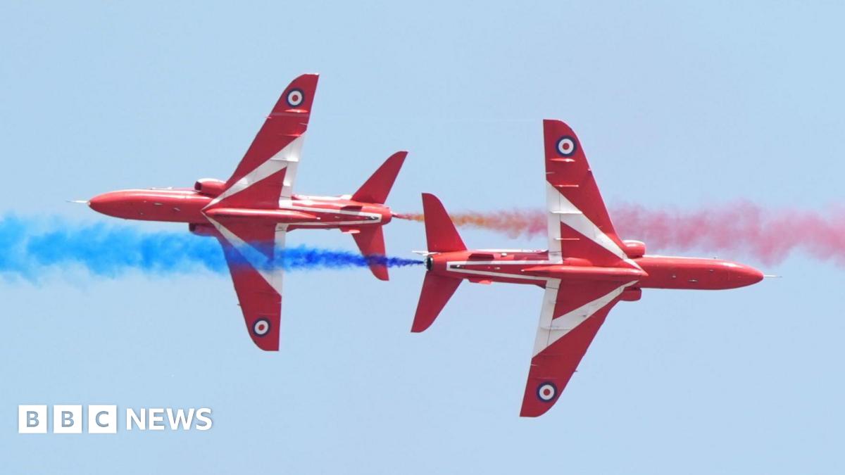 Two Red Arrows fighter planes, with red livery and a white triangle and red, white and blue bullseye symbol on their undercarriages, cross paths in the air while billowing out blue and red smoke
