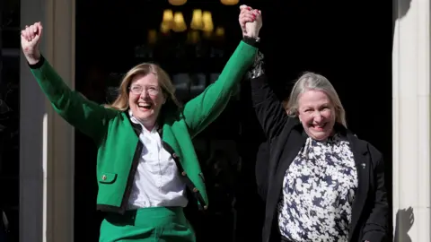 PA Media Two women celebrating outside the Supreme Court. The woman on the left is wearing a green suit with the jacket open over a white shirt. She has her hands in the air. Her left arm his being raised by the woman on the right, who has her right arm in the air. She is wearing a black jacket, which is open, over a blue and white floral patterned top.