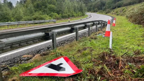 BBC The road is in a rural location with trees either side of the carriageway. A road sign showing a junction is on the ground in the foreground.