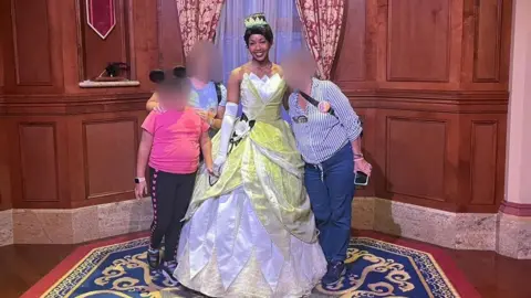Submitted Photo A woman in a blue-striped shirt, with her face blurred, stands next to Disney worker dressed as Princess Tiana in a yellow gown. The woman's daughter, face also blurred, stands on the other side of the princess. The daughter is wearing Mickey Mouse ears and a pink t-shirt