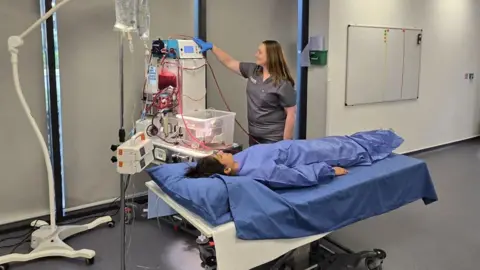 University of Oxford A woman wearing blue scrubs and lying on a hospital bed. A woman in grey scrubs is standing next to her, with her hand on a machine.
