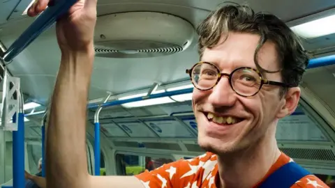 Joey Knock A man on the London underground smiles at the camera. He's holding his right arm up to hold onto the handrails and the photo looks like it was taken candidly. He's wearing an orange t-shirt with white stars and round tortoise-shell glasses. 