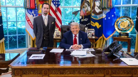 White House Trump sits behind desk in blue suit and red tie while Antoni stands in three-piece gray suit with a beige tie, white collar and watch chain in front of windows covered in colorful flags and banners