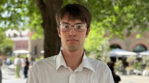 BBC Paddy Friend in a white shirt with a thin check pattern. He has brown hair and glasses.
