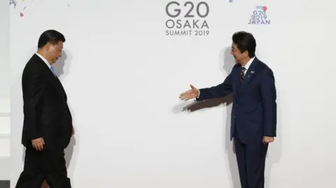 Getty Images Chinese President Xi Jinping looks down as he walks toward Japanese Prime Minister Shinzo Abe, who extends his hand with a smile