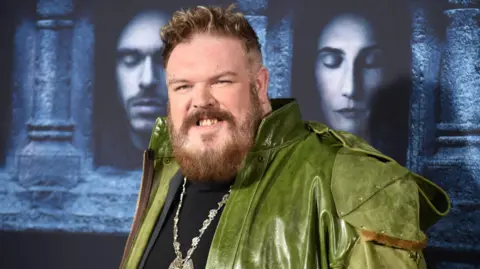 Getty Images Kristian Nairn wearing a green leather jacket and medallion standing in front of Game of Thrones graphics at the premiere for the sixth season at TCL Chinese Theatre on April 10, 2016 in Hollywood