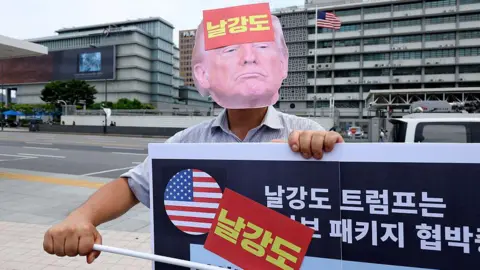 Getty Images A member of a civic group wearing a mask of US President Donald Trump stands during a protest against Trump's tariff policy near the US embassy on July 30, 2025 in Seoul, South Korea