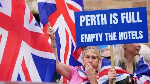 PA Media People demonstrating at an Abolish Asylum System protest outside the Radisson Hotel in Perth. Two blonde women are wrapped in Union flags and one holds a sign which reads "Perth is full empty the hotels"