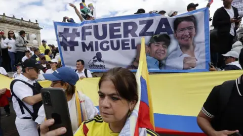 EPA People demonstrate with signs and flags during the "March of Silence" in Bogota, Colombia, 15 June 2025. A woman, wearing the shirt of Colombia's national football team, can be seen holding up a mobile phone. Behind her, people are carrying a banner reading "Strength, Miguel". 