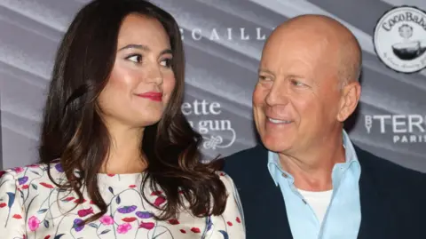 Getty Images Emma Heming Willis and Bruce Willis (Emma Heming is wearing a flowery dress and has long brown hair, Willis is wearing a light blue shirt and navy jacket)