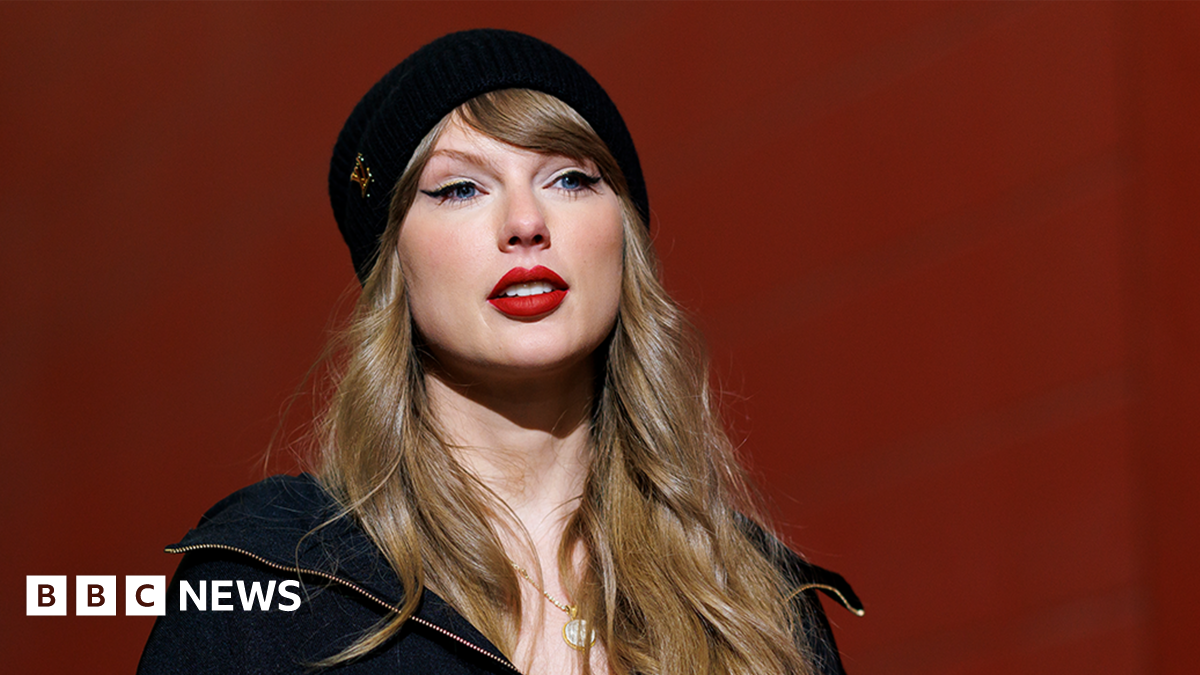 Taylor Swift smiling wearing a black beanie hat and black jacket.