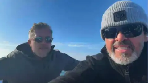 Robby West / BBC Two men sat next to each other on a boat. Matthew Parker (right) is wearing a grey beanie hat, black coat and black sunglasses. He is sat next to Mike Bates who is wearing a black coat and sunglasses. 