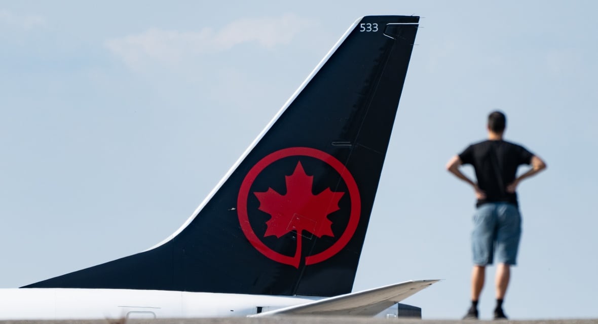 The tail of an airplane is seen, with a large red maple leaf on it. A blurry person stands in the foreground, their back to the camera. 