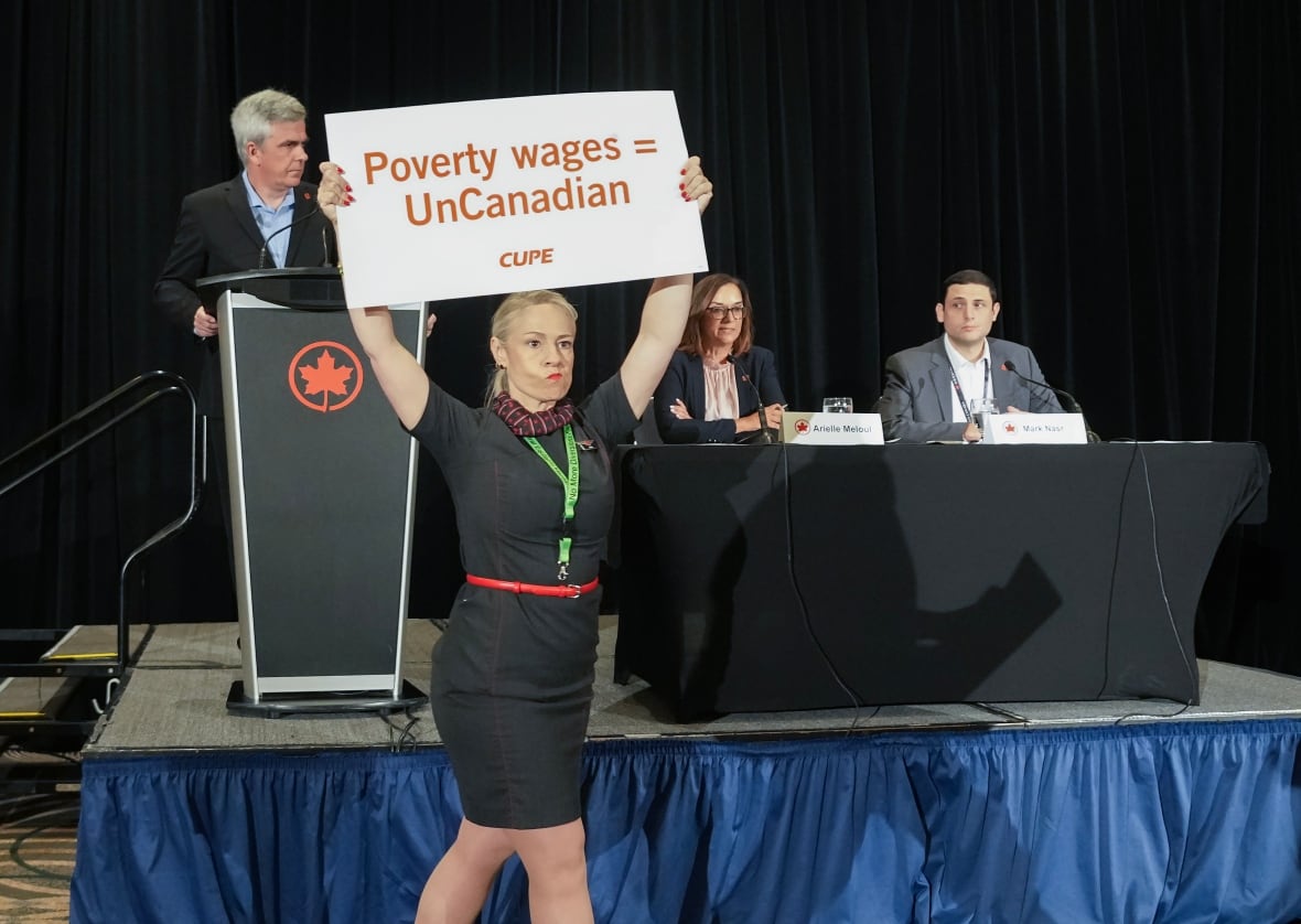 Air Canada officials at the head of a table inside a hotel ballroom, interrupted by protester holding up a sign that reads: Poverty wages = UnCanadian.