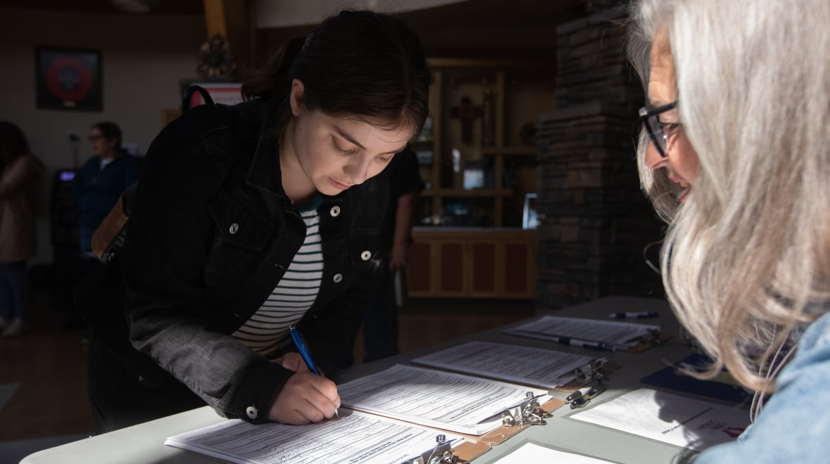 A young white woman, with dark hair tied back, is wearing a dark denim jacket and jeans. She leaning over a foldable table, signing a petition. An older woman, with long grey hair, is watching her.
