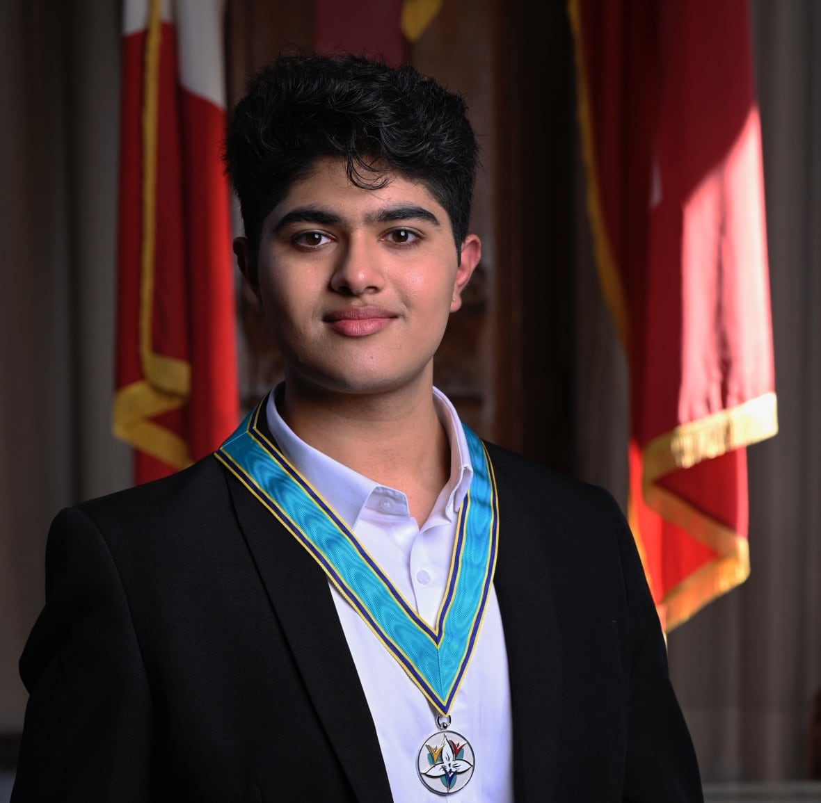 A portrait of a young man in a dark suit, wearing a medal on a blue ribbon around his neck, with several flags seen behind him. 