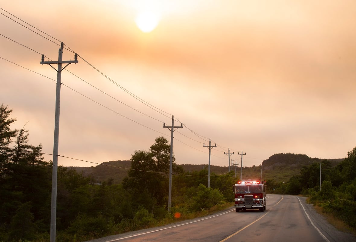 A lone fire truck drives along a forested road, against a smoky red sky.