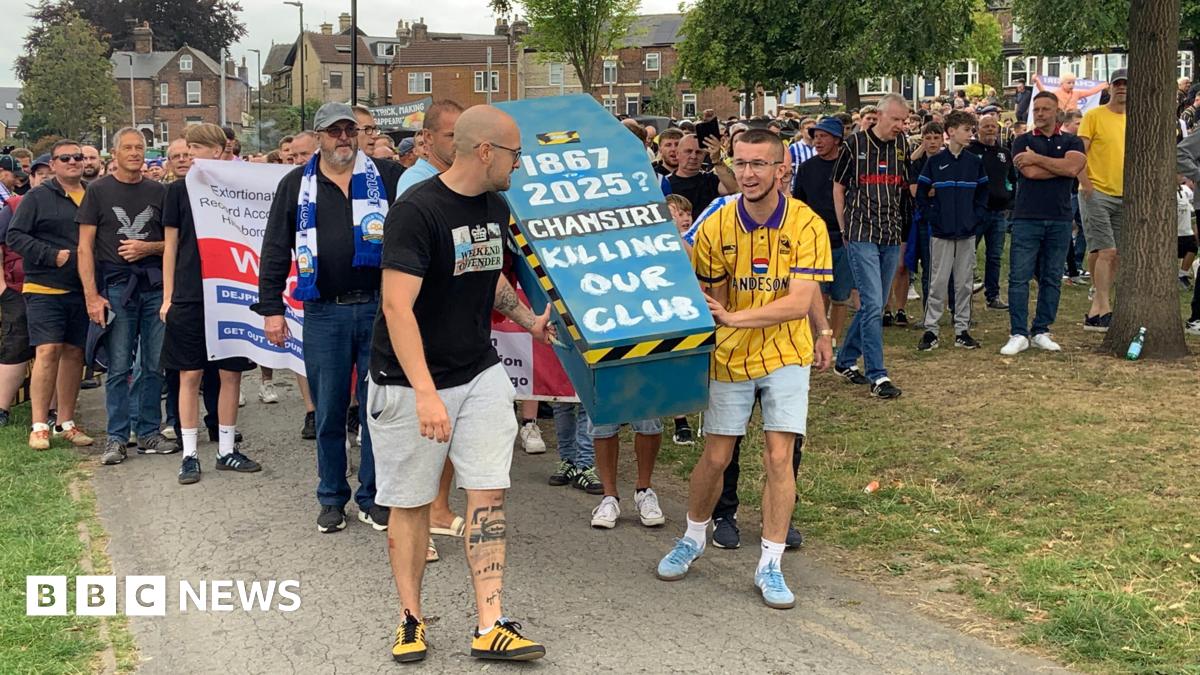 A large group of football fans are walking through a park. Some at the front of wearing football shirts in black and yellow and are carrying a mock coffin painted blue which says "Chansiri killing our club"