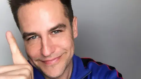 Anthony Kavanagh Anthony Kavanagh smiles as he holds an index finger up to celebrate one year of sobriety. He has dark hair and is wearing a blue top.